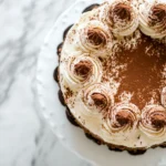 Oreo Tiramisu Cheesecake topped with cocoa powder and whipped cream on a white cake stand