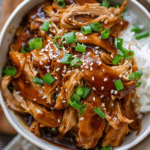 Crockpot Teriyaki Chicken shredded in teriyaki sauce, garnished with sesame seeds and green onions, served with steamed rice in a white bowl