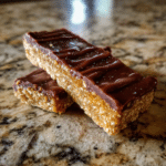 two homemade no-bake protein bars topped with a rich chocolate layer, stacked on a marble countertop