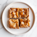 Maple Glazed Apple Blondies 6 maple glazed apple blondies with golden edges and soft apple-filled centers, arranged neatly on a white plate with shiny maple glaze